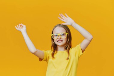 Sarı tişört ve gözlük takan neşeli kız, heyecan verici ellerini kaldırarak mutluluk teması için mükemmel bir zemin hazırlıyor.