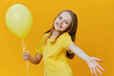 Sarı bir tişört giyen, elinde sarı bir balon tutan, parlak sarı bir arka plana gülümseyen, mutluluk ve kutlama duyguları uyandıran neşeli bir kız.