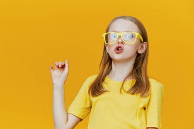 Neşeli genç bir kız sarı gözlük takar ve uyumlu bir tişört giyer. Parlak sarı bir arka plana karşı hoş yüz ifadesini sergiler. Mutluluk ve ilham verir.