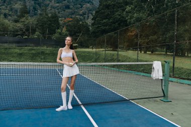 Şık bir tenis elbisesi giymiş enerjik genç bir kadın. Etrafı yemyeşil, spor, fitness ve sağlıklı görseller ile çevrili bir korta güven yayıyor.