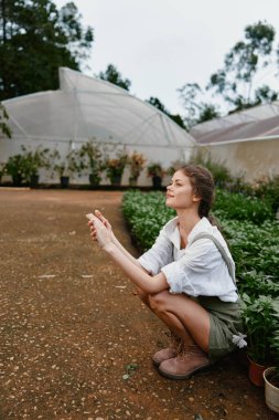Gür bir bahçede oturan genç bir kadın, günlük beyaz gömlek ve yeşil şort giymiş, doğaya neşe ve bağlantı saçan, çevre dostu yaşamı vurgulayan
