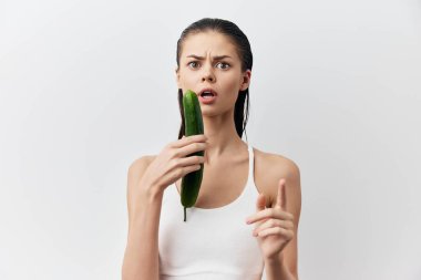 Şaşırmış bir kadın şok olmuş bir ifadeyle salatalık tutarken beyaz bir bluz giyer, beyaz bir arka plana yaslanır, sağlıklı beslenmenin mizahi yönünü yakalar ve...