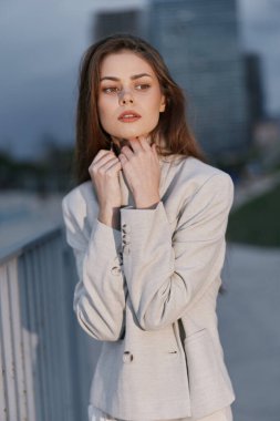 Açık bej ceketli düşünceli genç bir kadın, şehir arka planında, sessiz renklerle, derin düşüncelere dalmış bir şekilde poz veriyor, moda tarzında psikolojik ruh halini vurguluyor..
