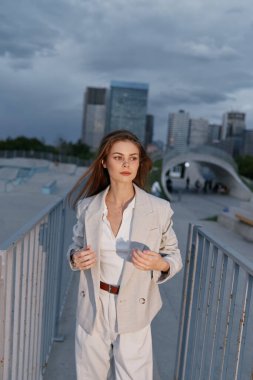 Kendine güvenen bej takım elbiseli bir iş kadını şehir siluetine karşı yürür. Modern bahar moda tarzında profesyonelliğini ve kararlılığını ifade eder..