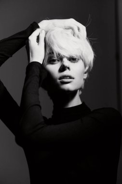 Kısa açık renk saçlı, siyah bluz giyen ve poz veren kendine güvenen bir kadın model. Yüksek kontrastlı ışıklandırma onun özelliklerini vurguluyor ve büyüleyici bir hava yaratıyor..