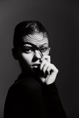 Modern modayı sergileyen bir kadının portresi, siyah bir elbise ve balık ağı başlığı takıyor, tek renkli bir ortamda gizem ve güven duygusu taşıyor..