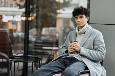 Bir kafede oturan, elinde bir fincan kahve, diğer elinde bir telefon olan şık genç adam pencereden dışarı bakıyor..