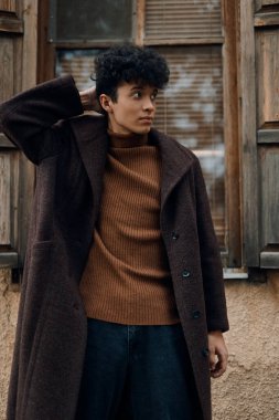 Kahverengi kazaklı ve ceketli şık genç adam önünde penceresi olan eski binanın önünde duruyor..