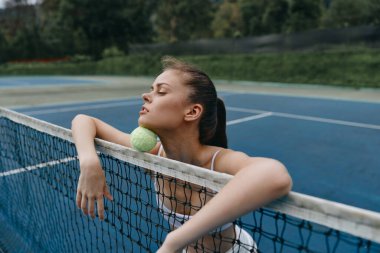 Tenis topuyla tenis sahasında oturan kendine güvenen genç bir kadın gevşeme sergiliyor ve yemyeşil yeşil bir tenis kortuyla çevrili canlı mavi bir tenis kortuna odaklanıyor.