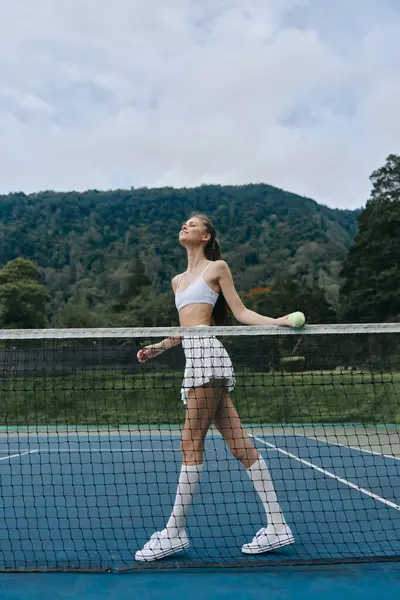 Enerjik genç tenisçi spor kıyafetleriyle canlı bir kortta güven içinde yürüyor, arka planda yemyeşil bir yeşillikle tenise olan neşe ve tutkuyu yansıtıyor.