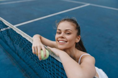 Mavi tenis kortunda tenis topuyla gezen neşeli genç bir kadın, yaz sporlarını, fitness 'i ve aktif bir yaşam tarzını temsil ediyor, neşe ve coşkuyu vurguluyor.