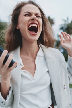 Hayal kırıklığına uğramış bir kadın dışarıda akıllı telefonuyla bağırıyor, ekonomik krizin duygularını beyan ediyor, beyaz gömlekli ve açık renk ceketli, dramatik bir yüz ifadesi var.