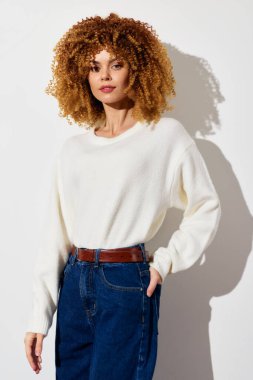 Moda mankeni, yüksek bel ve mavi kotla eşleştirilmiş rahat beyaz bir kazak sergiliyor. Açık arka plan güncel ama şık görünümünü arttırıyor, çağdaş modaya dikkat çekiyor.