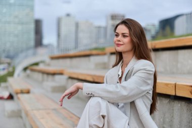 Açık renk takım elbiseli kendine güvenen bir kadın açık tribünlere rahatça oturur. Şehir manzarası arka planında, uzun kahverengi saçlar, sakin bir gülümseme, şehir modası, gündüz kuşağı.