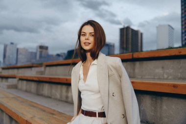 Bej ceketli ve beyaz bluzlu kendine güvenen bir kadın modern şehir manzaralı ahşap banklarda poz veriyor, sonbahar modasını ve iş tarzını gözler önüne seriyor.