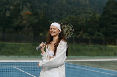 Neşeli genç bir kadın, açık havada, şık beyaz bir spor kıyafetiyle tenis oynamaktan hoşlanır, yemyeşil bir günün ortasında, parlak bir gülümsemeyle kendine güven yayar.