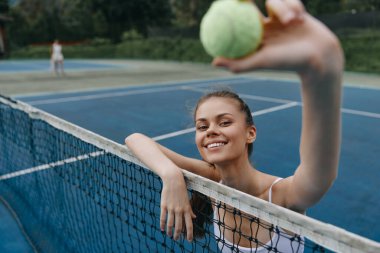 Enerjik genç bayan tenisçi mutlu bir şekilde ağların üzerinde canlı bir mavi kortta bir tenis topu tutuyor, spordaki coşku ve canlılığı somutlaştırıyor.