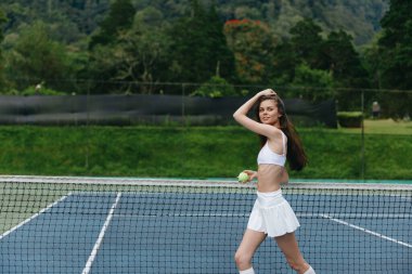 Beyaz spor kıyafetleri giyen enerjik genç bir kadın güneşli bir günde dışarıda tenis oynayarak sağlıklı bir yaşam tarzı ve sportmenliği teşvik ediyor.