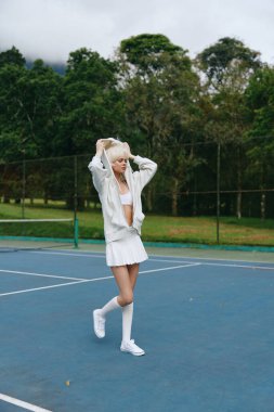 Beyaz tenis kıyafeti giymiş atletik bir kadın sahada poz veriyor, neşe saçıyor ve odaklanıyor spor, spor, spor ve açık hava aktiviteleri için mükemmel.