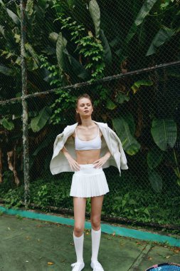 Beyaz spor kıyafetli kendine güvenen genç bir kadın tenis kortunda duruyor, canlı ve enerjik bir titreşim yayıyor yemyeşil yeşillikler arasında görüntü uygunluğu, tarzı ve doğayla olan bağlantısını yansıtıyor.