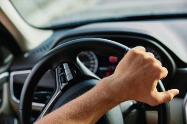 Direksiyondaki bir elin detaylı görüntüsü, bir aracın gösterge panelini ve iç unsurlarını gösteriyor.