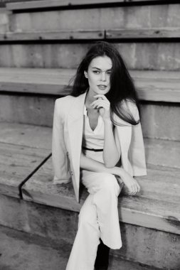 Beyaz takım elbiseli, kendine güvenen bir kadının siyah beyaz fotoğrafı. Merdivenlerde oturuyor. Düşünceli ve zarif görünüyor.
