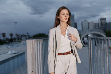 Şık takım elbiseli kendine güvenen bir kadın şehir ortamında duruyor, profesyonellik ve güç gösterisi yapıyor. Bulutlu gökyüzü onun güçlü varlığına dramatik bir dokunuş katıyor..