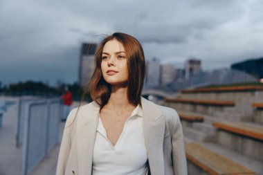 Şık bir iş elbisesi içinde kendine güvenen bir kadın açık havada şehir ortamında, hafif bir gülümseme ve sakin bir ifadeyle güç ve profesyonellik sunuyor..