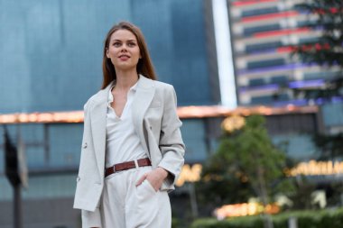 Bej renkli iş elbisesi giymiş kendine güvenen, modern şehir manzarası geçmişi olan, profesyonel tarzı olan, pozitif ruh hali olan, şirket başarısı için iş konsepti olan bir kadın.