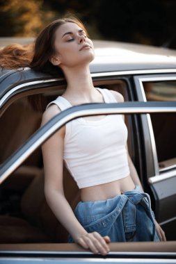 Yüzünde dingin bir ifade olan neşeli genç bir kadın, yaz özgürlüğünü somutlaştıran beyaz bir hasat bluzu ve kot giymiş bir arabanın kapısının dışındaki sıcak güneş ışığının tadını çıkarıyor..