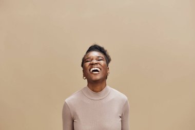 Açık renkli bir kazak giymiş neşeli bir zenci kadın geniş bir gülümseme sergiliyor. Mutluluğunu bej bir arka plana karşı sergiliyor. Bu görüntü, sevincin ve pozitifliğin özünü yakalıyor.