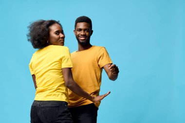 Neşeli bir Afrikalı çift, hareketler ve renkler yoluyla mutluluk ve romantizmi gösteren, canlı mavi bir arkaplan karşısında eşleşen sarı tişörtlerle neşeyle dans ediyor.