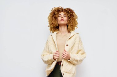 Süslü bir kadın model, krem rengi büyük bir süveter ve bej bir bluz giyiyor, minimalist beyaz bir arkaplanda poz veriyor kıvırcık saçları şık kıyafete dinamik bir dokunuş katıyor.