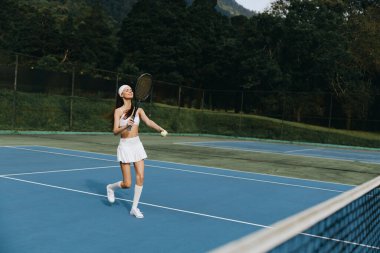 Beyaz spor kıyafeti giymiş neşeli bayan tenisçi, açık mavi bir kortta oynuyor, atletizm ve açık hava sporlarının mutluluğunu canlı bir doğal ortamda sergiliyor.