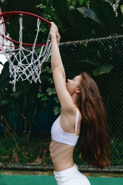 Enerjik genç basketbolcu, dinamik bir atmosferi vurgulayan yemyeşil bir arka planla şık bir spor kıyafetle neşe ve zafer ifade ediyor.
