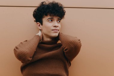 Kıvırcık saçlı, kahverengi kazak giyen, elleri başının üzerinde duvara yaslanmış ve kameraya bakan genç bir adamın moda portresi..