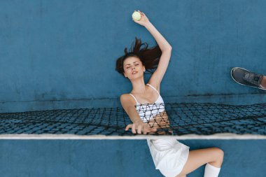 Spor kıyafetleri içinde atletik genç bir kadın tenis oynar, parlak mavi bir zemin üzerinde heyecan ve hareket ifade eder, spor ve sağlıklı yaşam temaları için mükemmeldir.