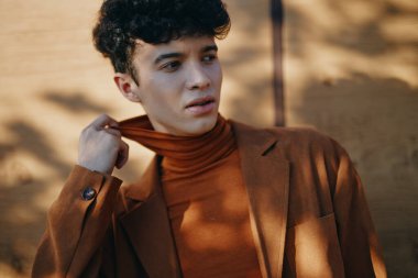 Kahverengi balıkçı yaka ve ceket giyen şık genç bir erkek, yumuşak gölgeli modern bir görünüm sergiliyor.