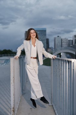 Kendine güvenen bej takım elbiseli bir kadın bulutlu gökyüzünün altında şehir merkezinde yürüyor, gülümsüyor, modern moda tarzını ve profesyonel zarafetini sergiliyor.