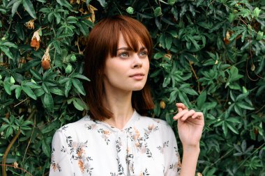 Canlı kızıl saçlı kadın zarif bir şekilde çiçekli yeşil çalıların önünde duruyor.