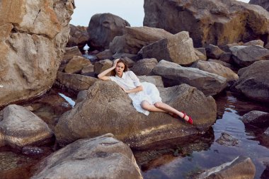 Huzurlu sularda kayaların üzerinde oturan, huzurlu manzarayı seyreden, beyaz elbiseli sakin bir kadın.
