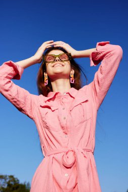 Pembe elbiseli ve şık aksesuarlı neşeli genç bir kadın güneşli açık hava atmosferini kucaklıyor açık mavi gökyüzüne karşı yaz modası için mükemmel
