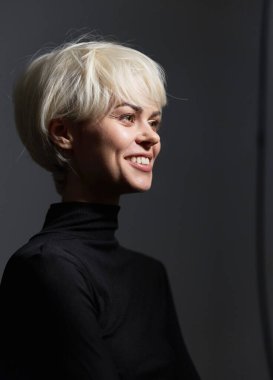 Neşeli bir bayan model, parlak bir gülümseme, modern saç stili ve nötr bir arkaplan karşısında minimalist moda sergiliyor. Duygulara ve bireyselliğe vurgu.