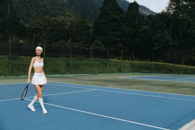 Beyaz tenis kıyafetleri içinde atletik bir kız mavi bir tenis kortunda kendine güvenen bir yürüyüşün keyfini çıkarıyor, canlı bir spor yaşam tarzını manzaralı bir dağ manzarasına karşı sergiliyor.