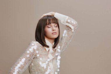 Genç bir kadın, omuzları pullu, siyah saçlı, tarafsız bir arkaplanda duran, kameraya özgüvenle bakan, çağdaş moda gösteren