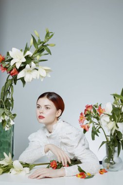 Beyaz bir bluz içinde zarif bir genç kadın, canlı çiçeklerle düşünceli bir şekilde poz vermiş, güzelliğin, sükunetin ve çiçeklerin özünü modern bir ortamda yakalıyor.