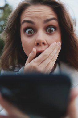 Genç, şaşkın, gözleri açık ve ağzı açık bir kadın akıllı telefona, günlük kıyafete, doğal arka plana, psikolojik konsepte bakarken duygusal tepki veriyor.
