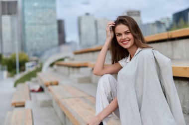 Hafif takım elbiseli gülümseyen kadın şehir manzarası arka planında şehir tribünlerinde poz veriyor, günlük moda kıyafeti, doğal ışık ve bahar sezonunda pozitif özgüven duygusu.