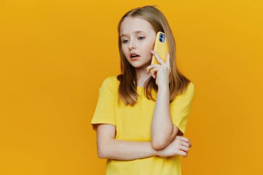 Bu görüntüde sarı tişörtlü genç bir kız canlı sarı bir arkaplan önünde sarı bir telefonla konuşuyor, şaşırdığını ve merakını dile getiriyor.