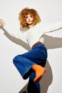 Moda mankeni, kıvırcık saçlı, temiz beyaz bir fonda poz veren, rahat formalı bir süveter ve kot giyen, parlak turuncu çoraplı parlak bir popo ekleyen, eğlenceli bir görünüm için.